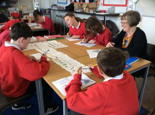 Primary school aged children in a classroom setting, sitting at desks completing an interactive card activity. An adult is sat supporting the children and smiling.