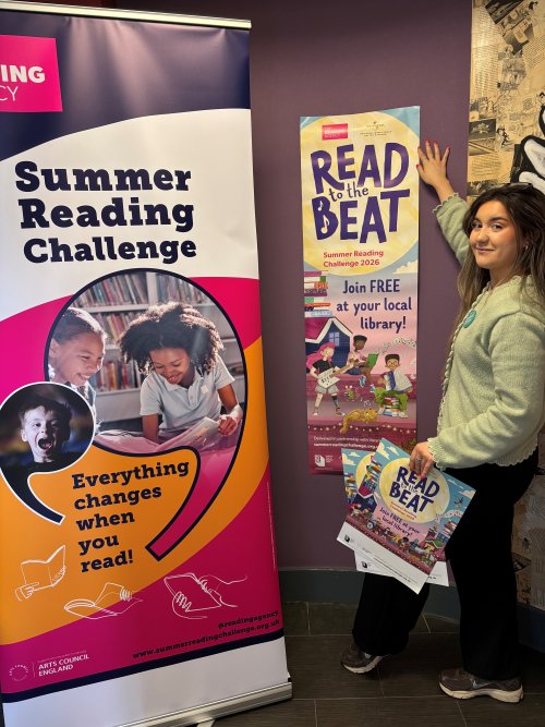 A woman standing next to a 'Read the Beat' Poster and a 'Summer Reading Challenge' banner.