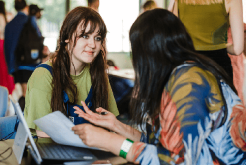 A teenage girl and an adult woman engaged in conversation across a table, with a laptop and notes between them at a mentoring event.