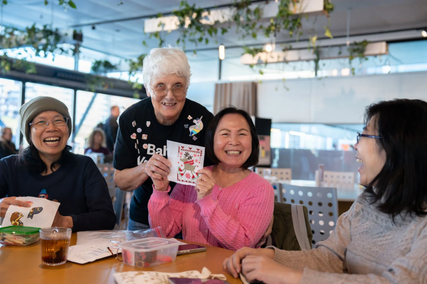 A person at a table with friends hold up her sewing and smiles at the camera    