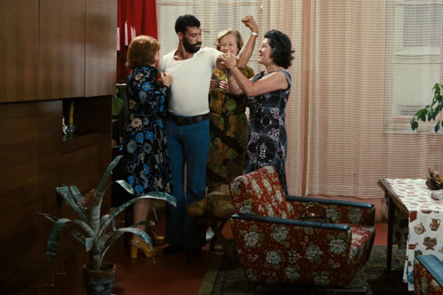 A man stands flexing his arm muscle whilst surrounded by a group of woman. 
