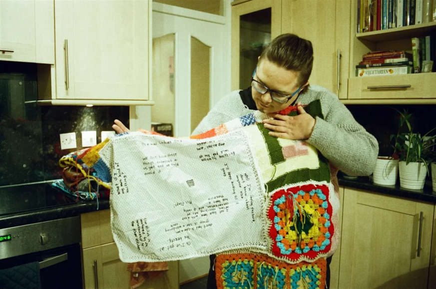 colour photograph, interior. A Drag King with glasses and a knitted jumper looks down at his knitted blanket which he holds open across his chest. He stands in a kitchen.