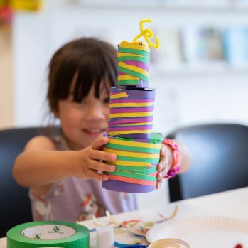 A child sits at a table in a brighly lit room. She holds up and presents a handmade sculpture, a tower of brightly coloured paper, pipe cleaners and  tape . On the table there are rolls of green tape and glue sticks.