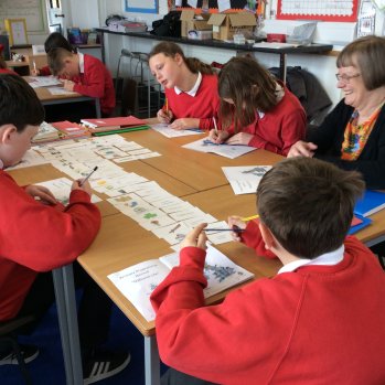 Primary school aged children in a classroom setting, sitting at desks completing an interactive card activity. An adult is sat supporting the children and smiling.