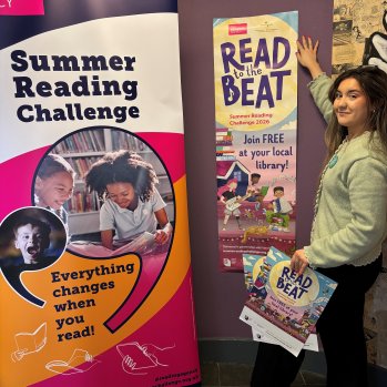 A woman standing next to a 'Read the Beat' Poster and a 'Summer Reading Challenge' banner.