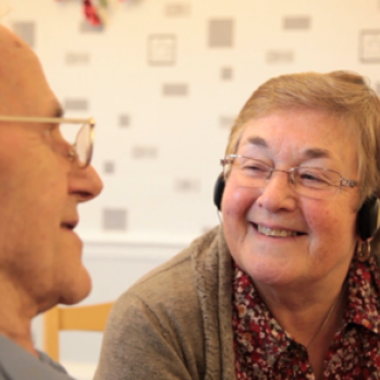 Two older people wearing headphones and smiling at each other.