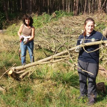 Two ladies in a forest clearing branches and smiling at the camera.